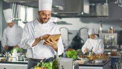 Famous Chef Uses Tablet Computer for Recipes While Working in a Modern Kitchen. His Help Work in the Background.