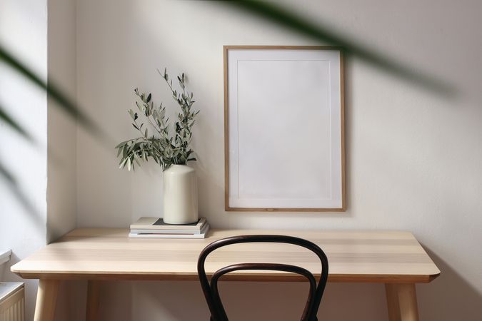 Empty vertical picture frame mockup. Wooden desk, table. Vase with olive branches, magazines. Elegant working space, home office. Summer interior design. Beige walls. Blurred palm leaf overlay.
