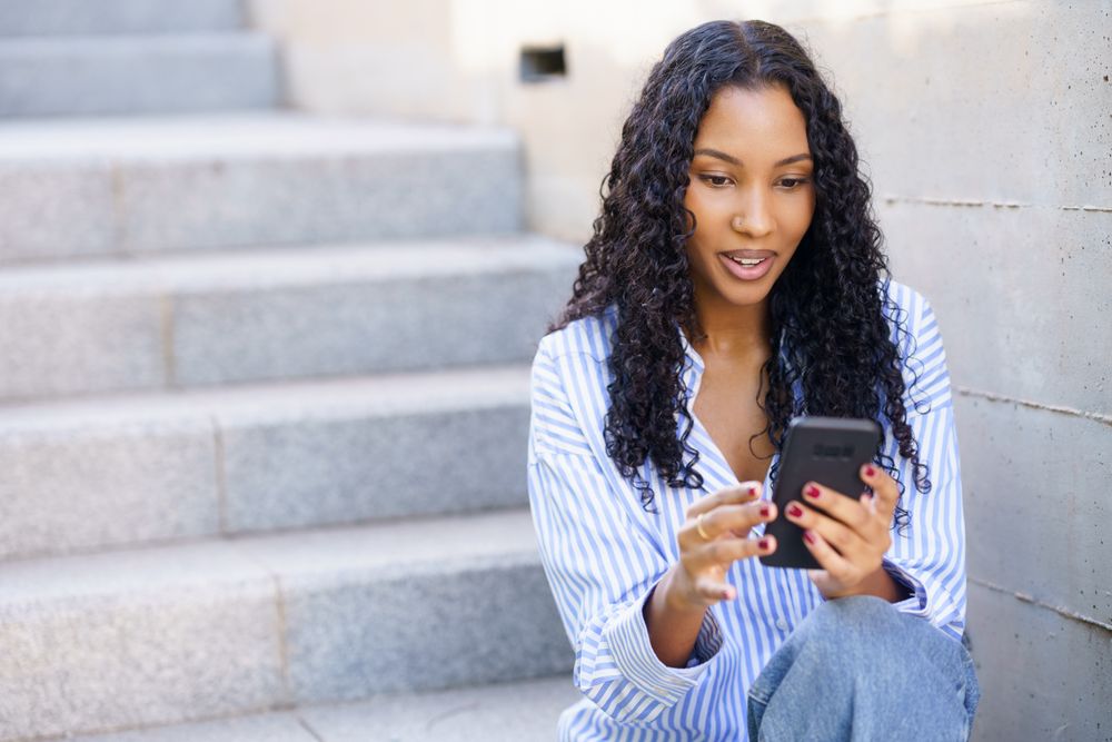 A young woman is engaged with her smartphone while sitting on outdoor steps, smiling happily