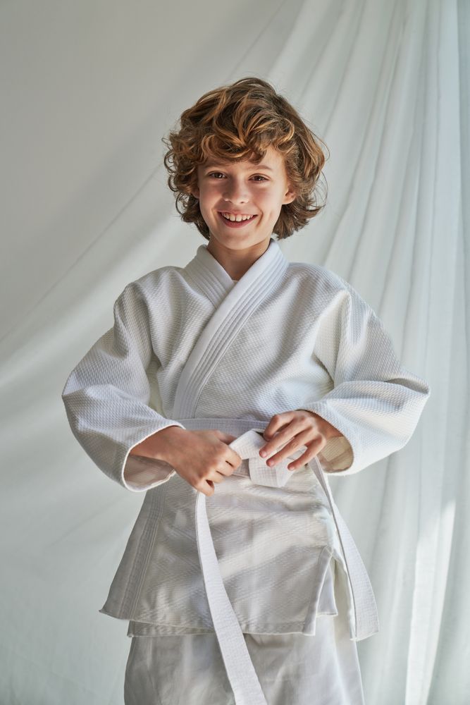 Positive preteen karate kid in kimono standing near curtain