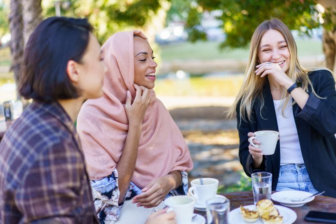 Three smiling women laughing while enjoying coffee and baked goods on a beautiful sunny day