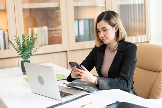 Woman looking at phone while sitting at her desk with laptop