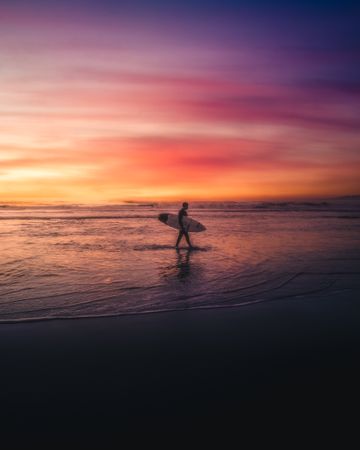 Silhouette of man holding surfboard and walking on beach during sunset