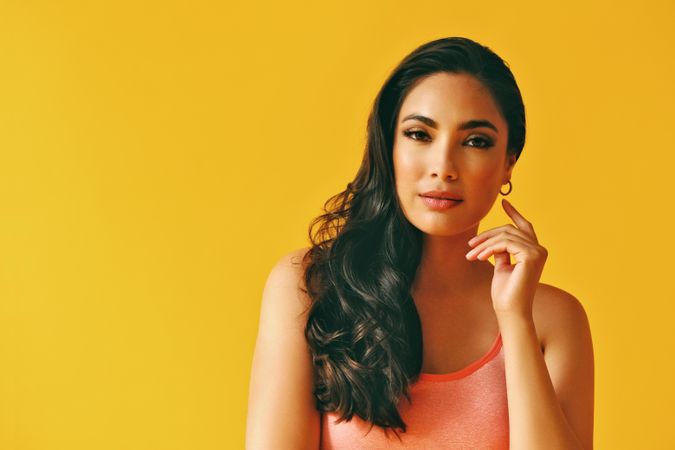 Headshot of calm Hispanic woman looking at camera in yellow room, with hand up to her cheek