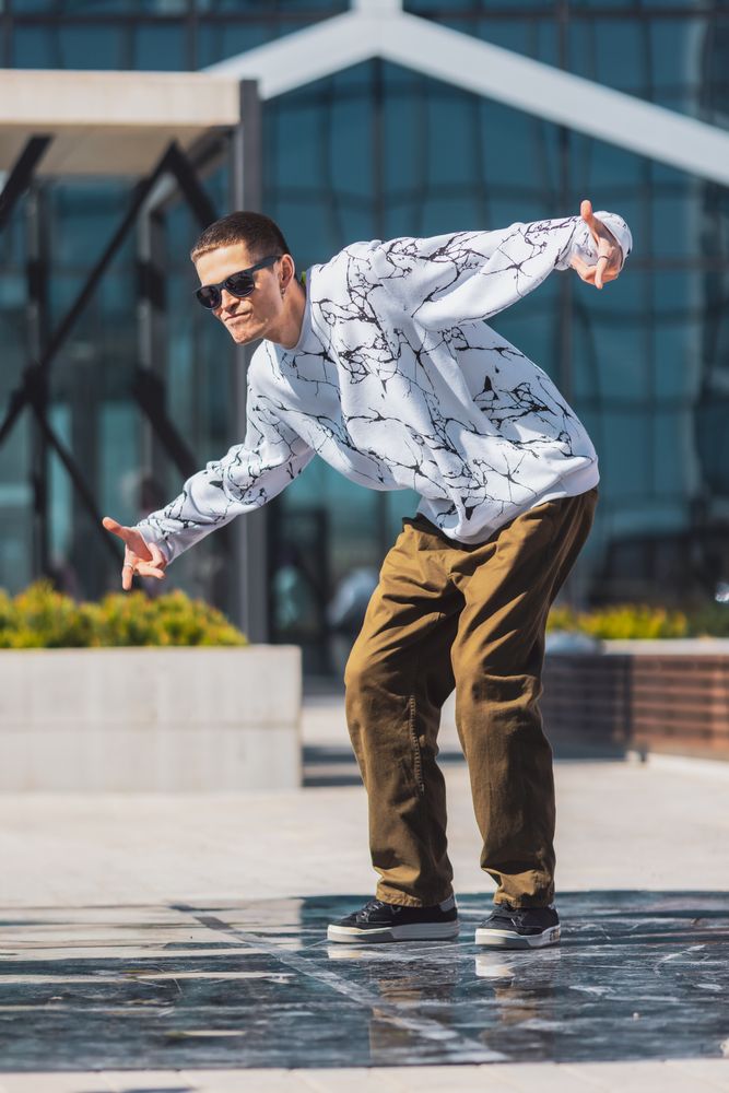 Young Man In Stylish White Marble Pattern Sweatshirt And Brown Pants Performing A Dynamic Dance Move In An Urban Setting With Modern Glass Buildings In The Background.