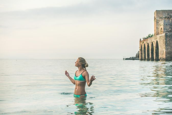 Woman in bikini standing in the calm sea with her eyes closed and hands raised