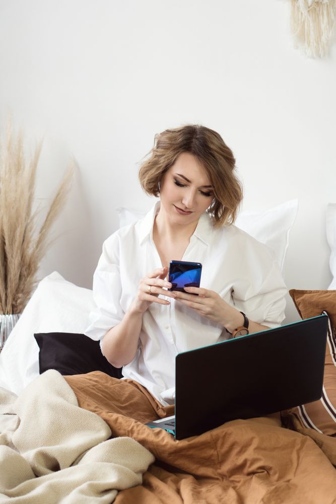 Woman Sitting In Bed Spending Time In Bedroom With Phone