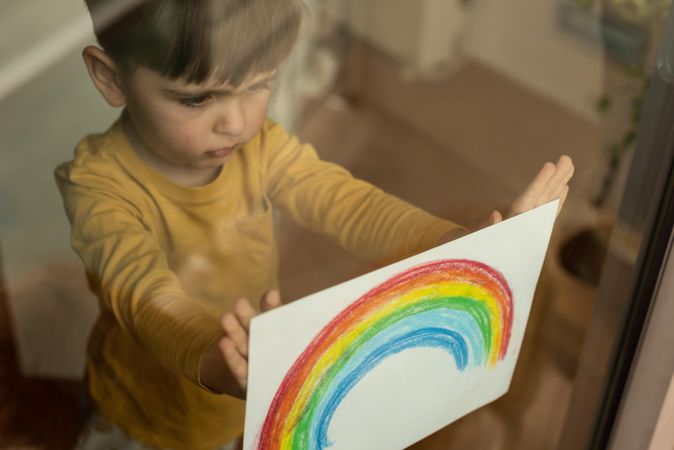 Young boy holding rainbow drawing to the window