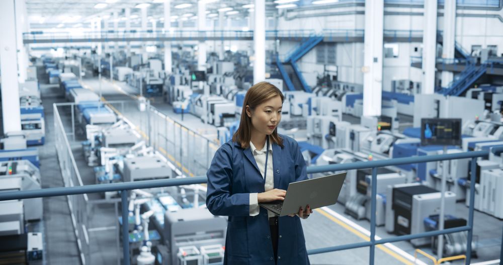 Young Asian Female Engineer Standing on a Platform, Using Laptop Computer at an Electronics Factory. Machines are Undergoing Maintenance, Specialist Monitoring the Progress Through Online Software