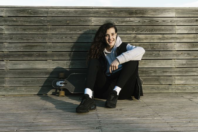 Teenage girl sitting on a longboard, leaning against a wooded wall