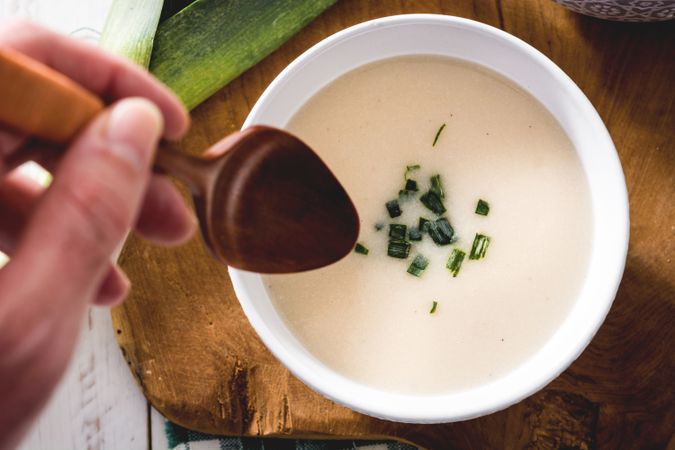 crop hand with spoon and tasty Vichyssoise soup on wooden table with leek