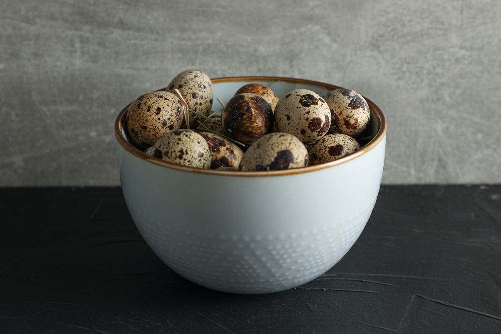 Quail eggs in bowl on dark table against grey background, space for text