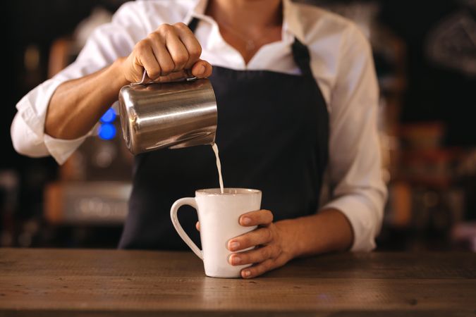 Woman wearing apron pouring milk into a cup of coffee, making espresso