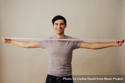 Smiling Hispanic male holding measuring tape wide between both arms in beige studio shoot 0L7Xrb