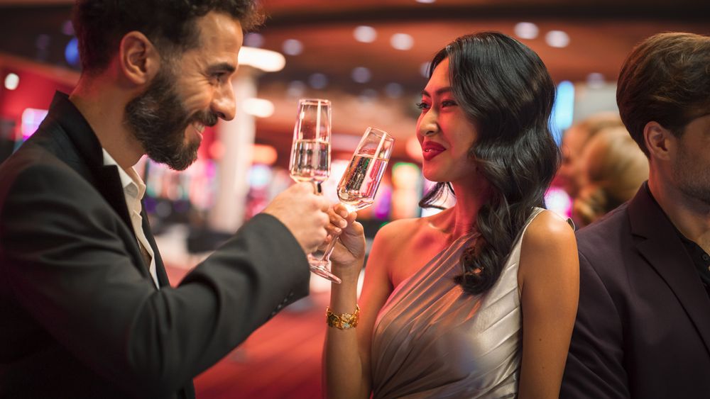 Multiethnic Couple Having a Fun Conversation at a Casino while Drinking Champagne and Gambling at a Roulette Table.