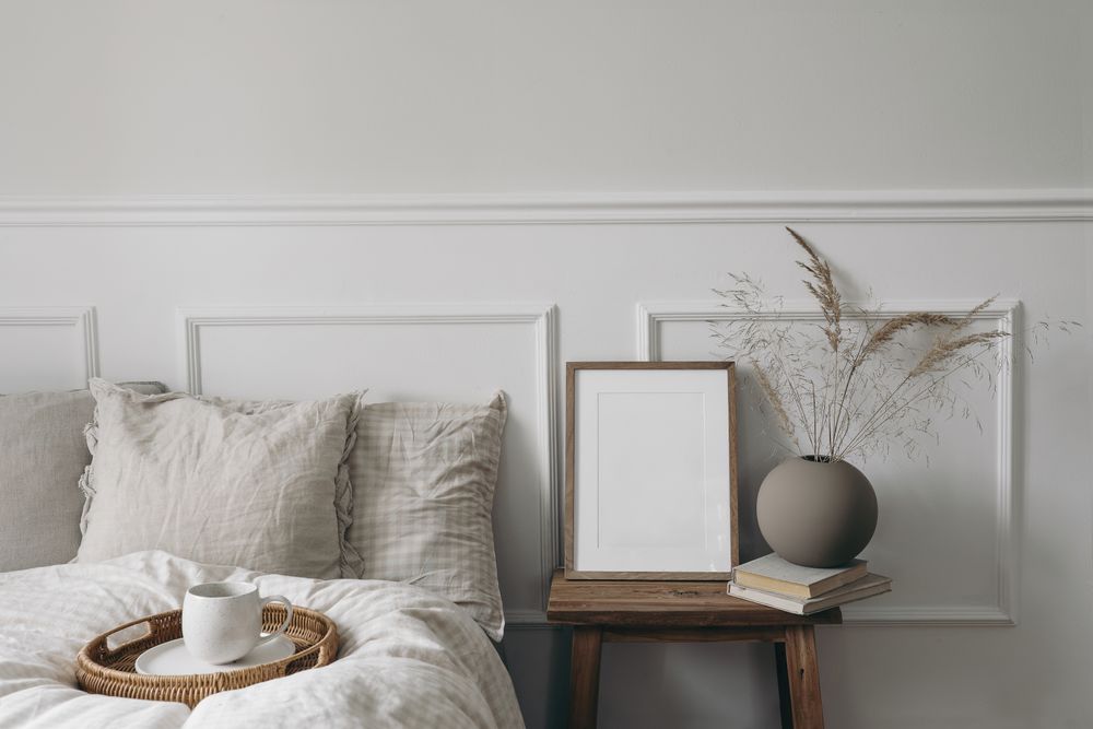 Bedside table with mockup frame and vase next to comfortable bed with tray of coffee