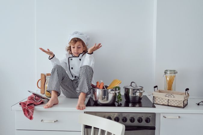 Bewildered preteen boy in chef costume sitting on kitchen counte