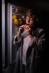 Pleased boy eating near opened fridge in dark kitchen