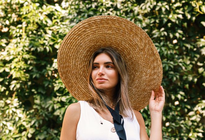 Woman adjusting straw hat standing in front of a bush