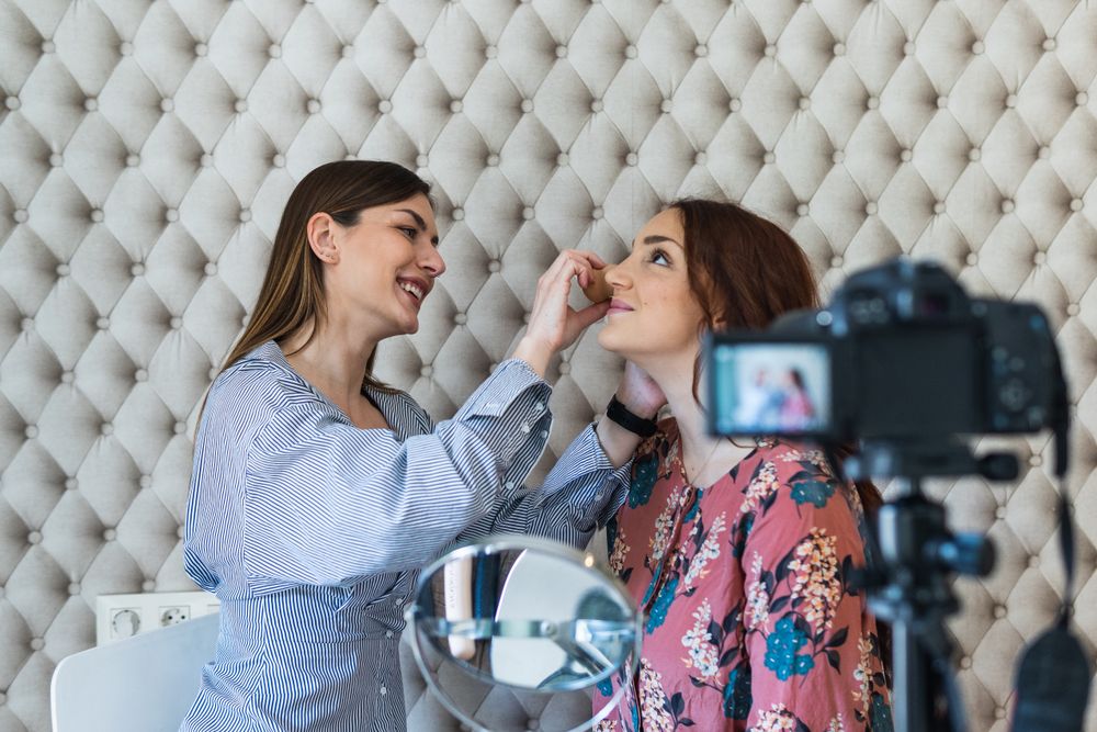 Cheerful woman applying makeup on model and recording video