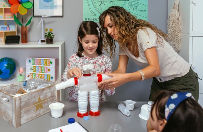 Teacher helping to girl make recycled toy robot with plastic packages
