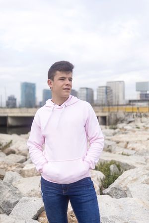 Male teenager standing against cityscape with hands in pocket on breakwaters while looking away