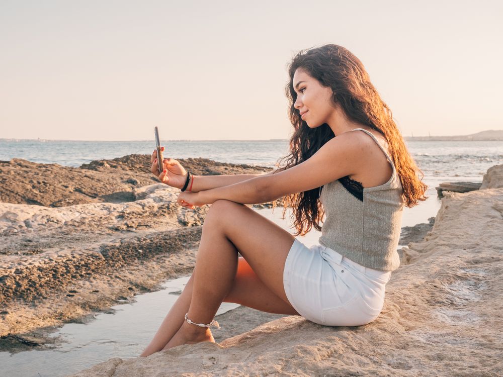 Dreamy trendy young woman texting on beach