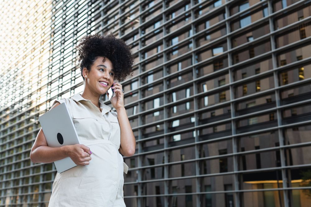 Smiling Black pregnant woman talking on smartphone and carrying a laptop