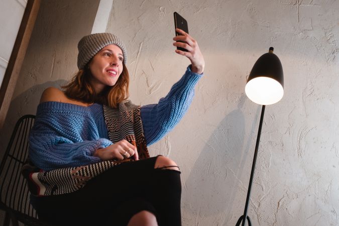 Woman in sweater and hat taking selfie and sitting near wall