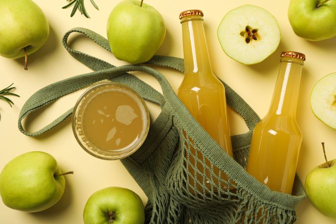Bottles and glass with cider, green apples and bag on beige background, top view