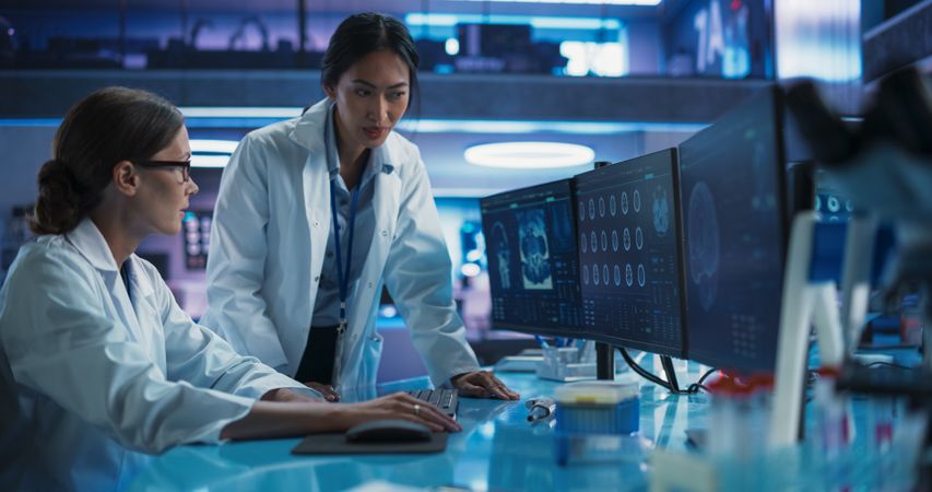 Medical Hospital Research Laboratory:  Female Scientists Talking And Using Desktop Computer With CT Scans Of Human Brain.