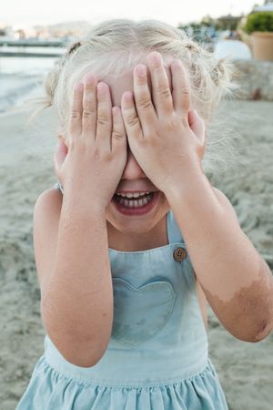 Shy adorable girl hiding face with hands and smiling on sandy sh