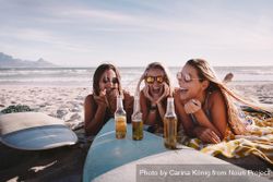 Group of young women sitting in the sand with a surfboard and beer 25n6Q5