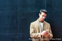 Man in fall coat leaning back on dark outdoor wall while texting 4jVRAX
