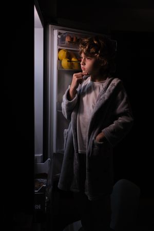 Thoughtful boy near opened refrigerator in dark kitchen