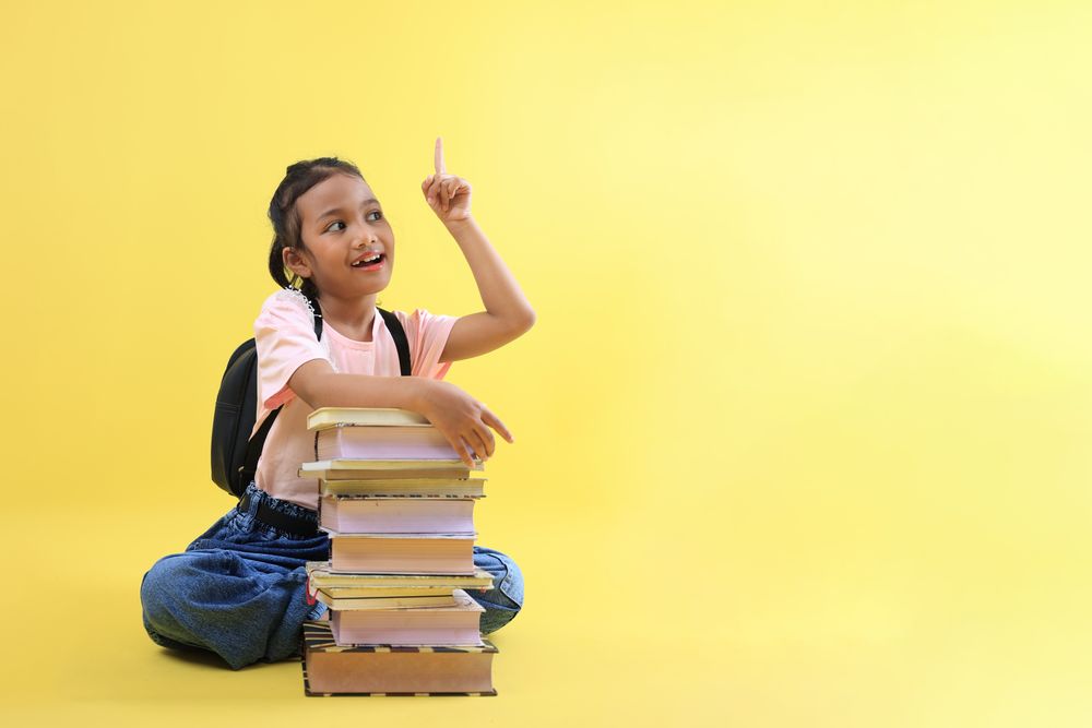 Little Asian Girl Sitting Wearing Casual Clothes and Jeans, Pointing to the Top, Her Hand on Stacked Book.