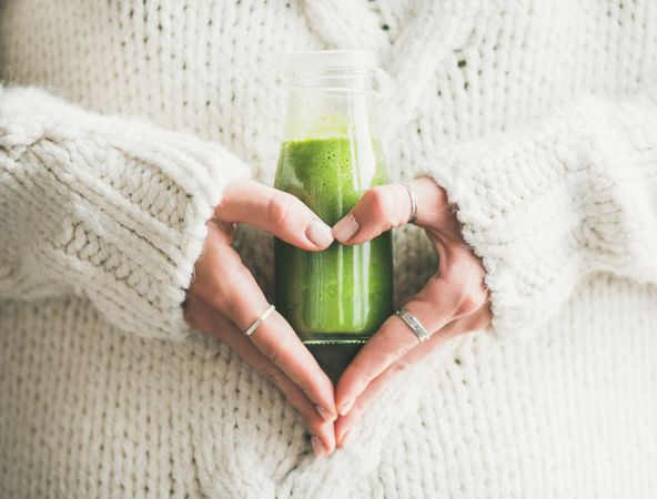 Woman in sweater,  making heart shape with hands, with green smoothie horizontal composition