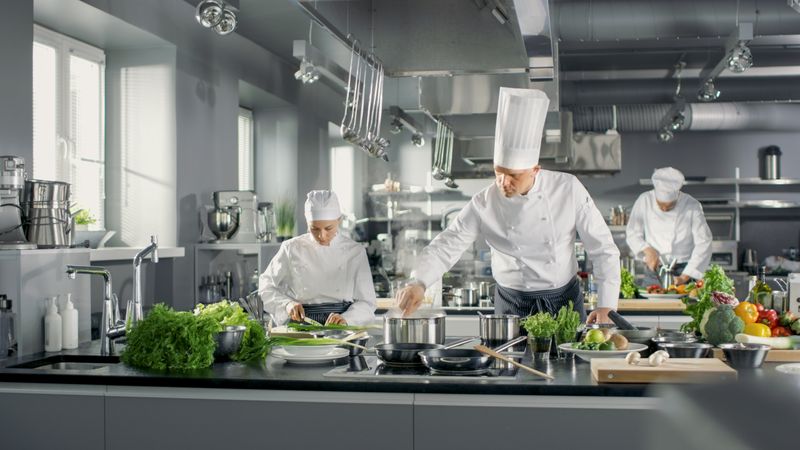 Famous Chef Works in a Big Restaurant Kitchen with His Help. Kitchen is Full of Food, Vegetables and Boiling Dishes.