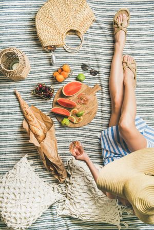 Woman on picnic blanket with  thatched bag with glass of rose, baguette, sliced watermelon and figs