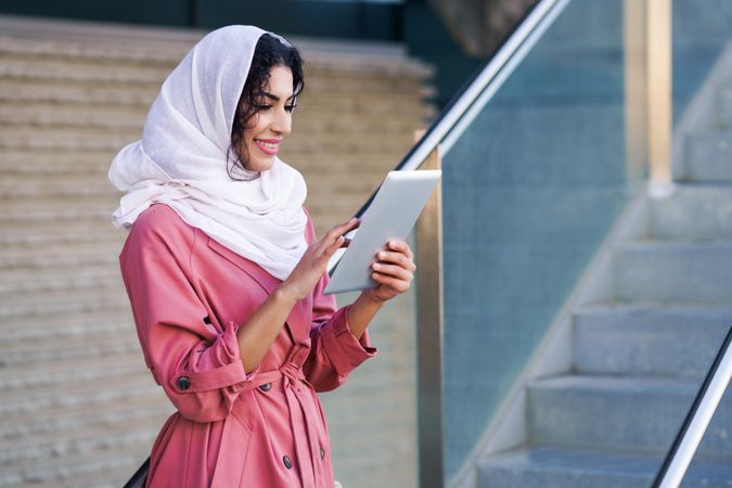 Happy female in headscarf and pink trench coat with her digital tablet near staircase