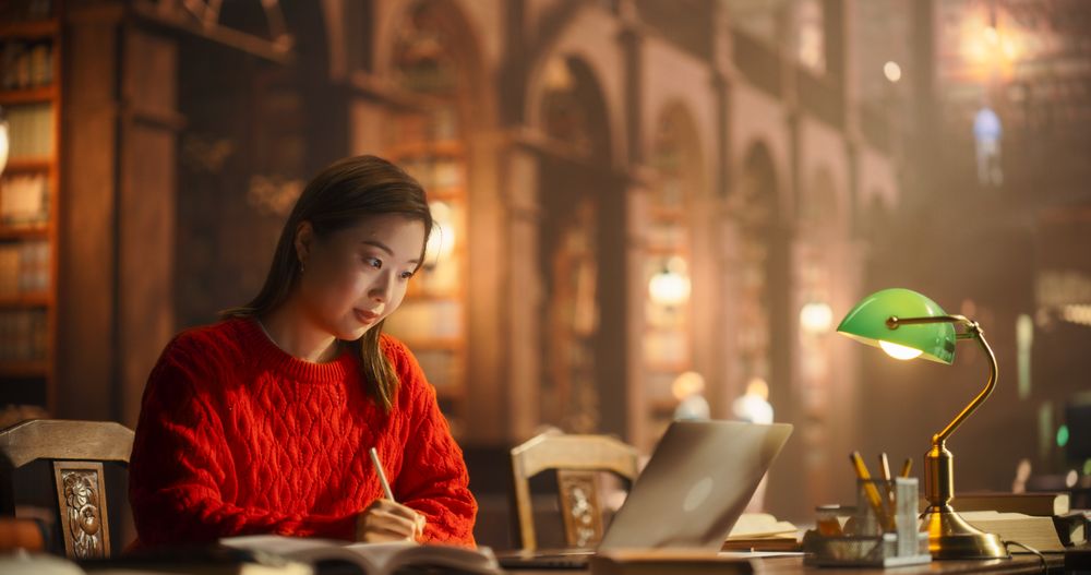 Asian Female Student Engaged in Studying With Books and Laptop in Classical Library Setting. Young Woman Focused on E-Learning