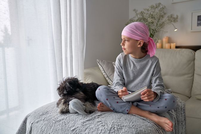 Pensive boy in pink headscarf writing in notepad near dog
