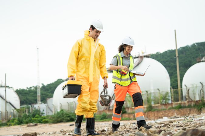 Workers in safety gear walking to survey at site near refinery storage tanks, industry facility, Hazardous environment, scientist holding toolbox, woman with laptop