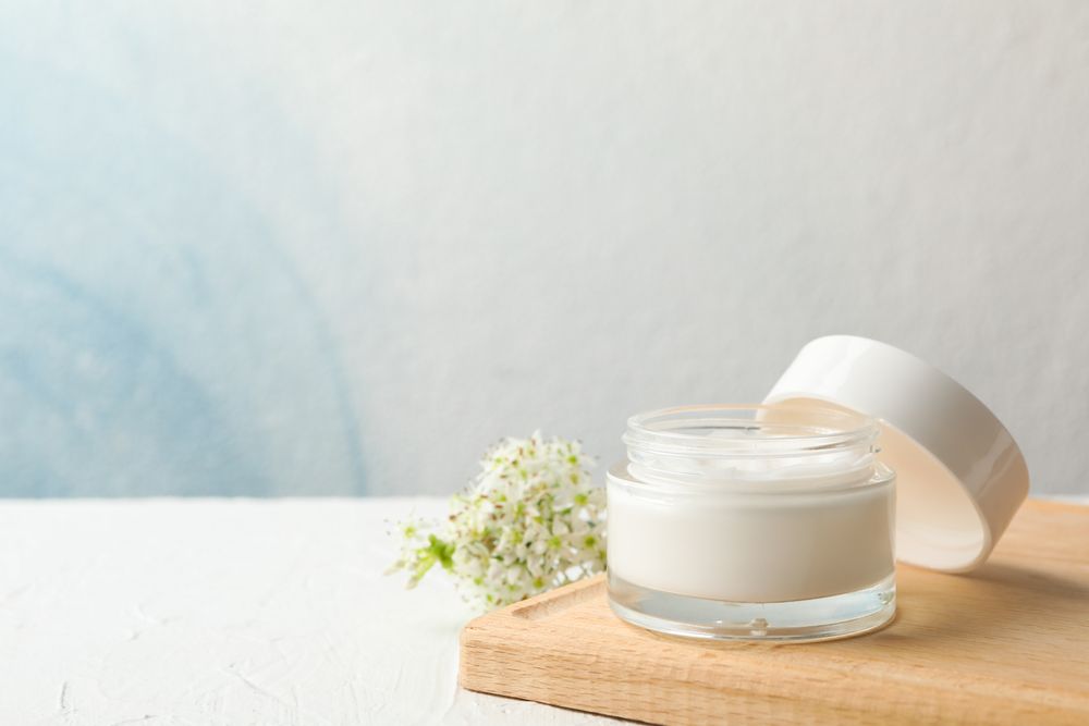 Jar with cream, allium flowers and board on neutral background, copy space