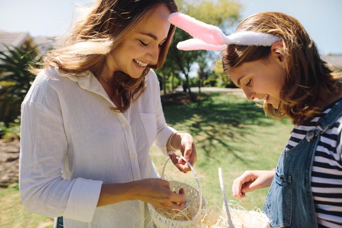 Smiling woman putting easter egg sweets in baskets standing outdoors with her daughter
