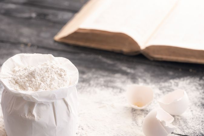 Open bag of wheat flour on rustic wooden table covered with flour, egg shells and recipe book