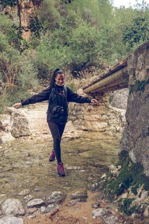 Happy woman crossing shallow river while hiking in rocky terrain