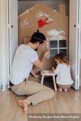 Back view of father and young daughter building a small house out of cardboard at home 0ypEWb