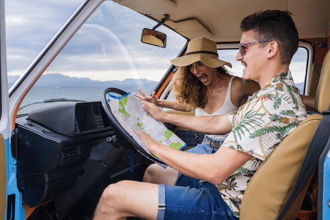 Touristic man exploring map with help of long haired woman in hat in front seat of car