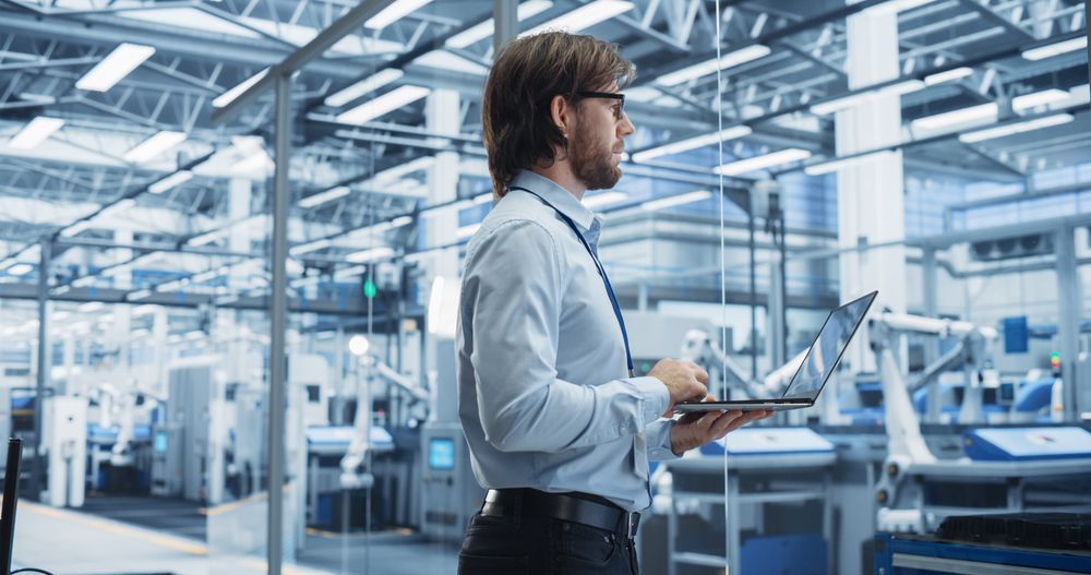 Experienced Male Engineer Standing in Office, Using Laptop Computer and Overlooking Production at a Modern Automated Electronics Assembly Line with AI Support. Factory Executive at Work